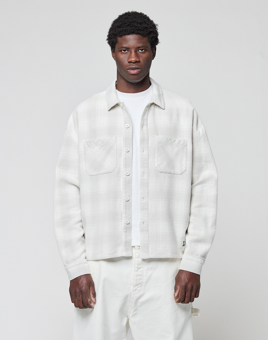 A man with short, curly hair and a neutral expression stands facing forward, wearing a LIVE FAST DIE YOUNG Vintage Flannel in an oversize fit over a white T-shirt and white pants against a plain light gray background.