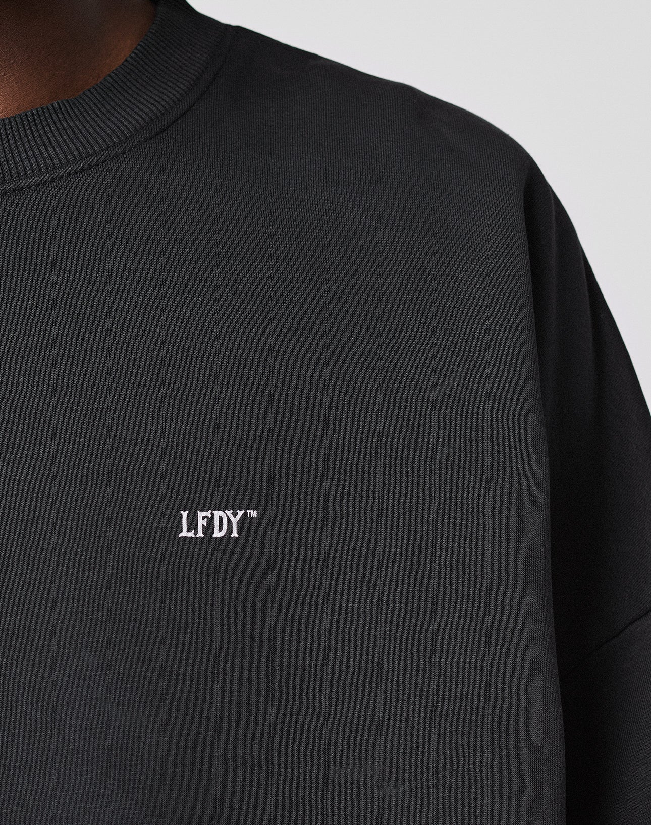 A close-up shows a person in the LIVE FAST DIE YOUNG Basic Evidence Sweater, an oversize fit black sweatshirt featuring small white embroidered LFDY™ text on the chest. Only part of their neck and shoulder is visible.