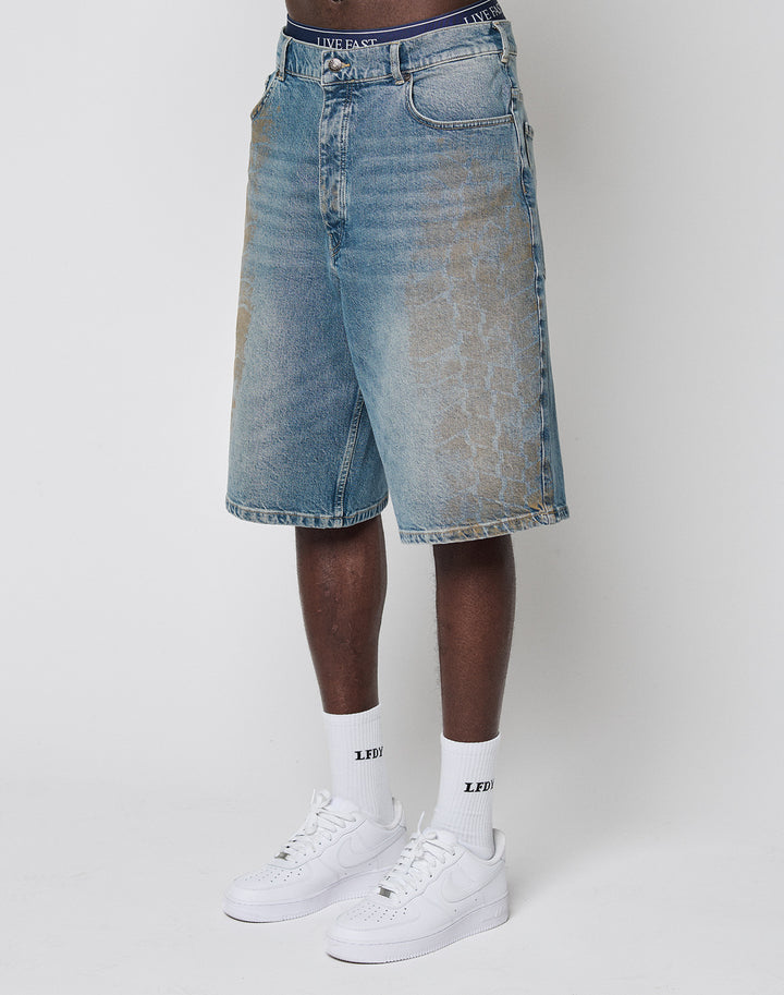 A person wears faded, baggy fit LFDY Tire Tracks Jorts by LIVE FAST DIE YOUNG, knee-length denim shorts, paired with white socks featuring LEDI text and white sneakers. Only the lower body is visible against a plain white background.
