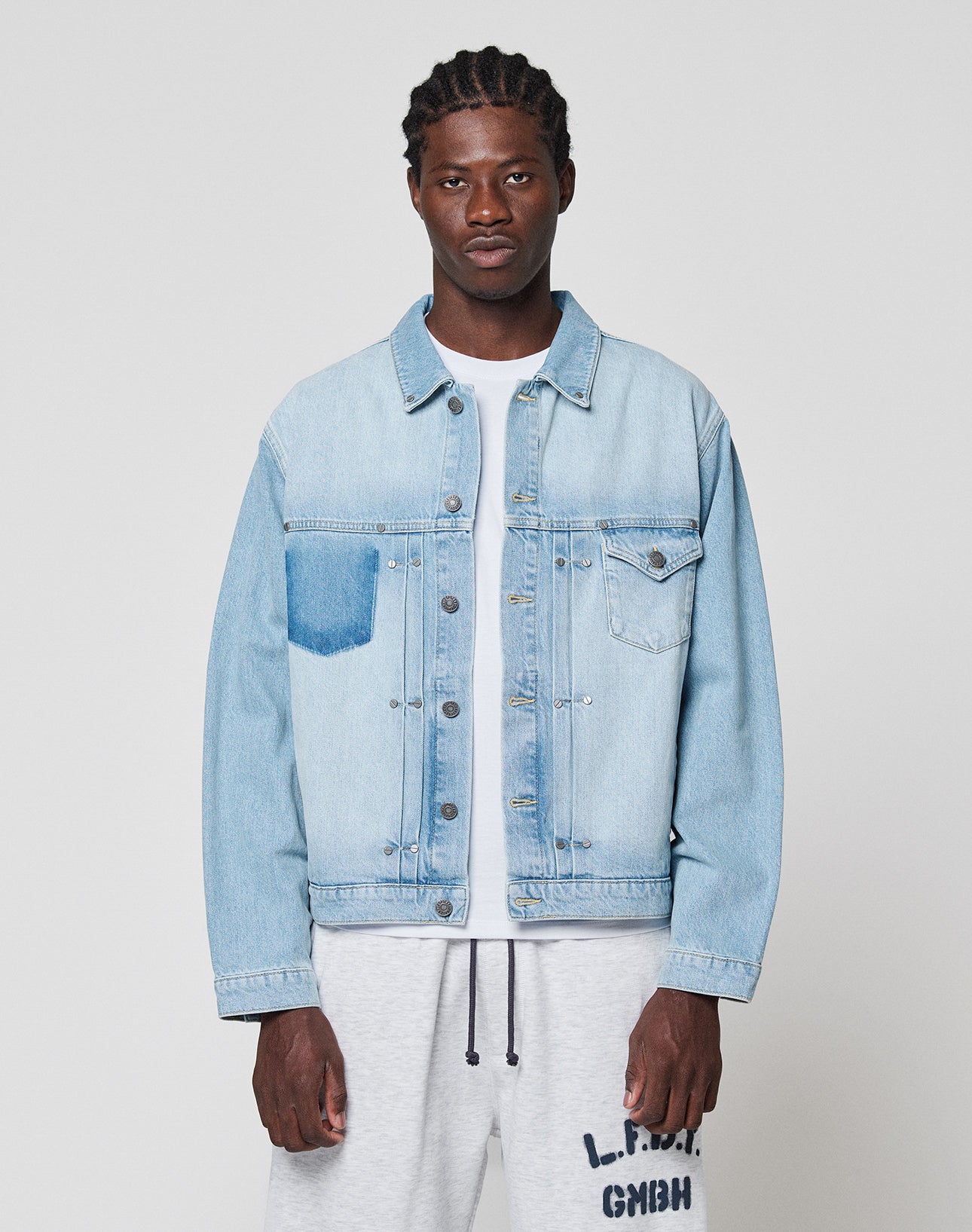 A man faces the camera wearing a LIVE FAST DIE YOUNG LFDY Trucker Jacket in light blue denim with one dark wash pocket, crafted from Baumwolle in Regular Fit, paired with a white T-shirt and light gray sweatpants with black text. Background is plain and light.