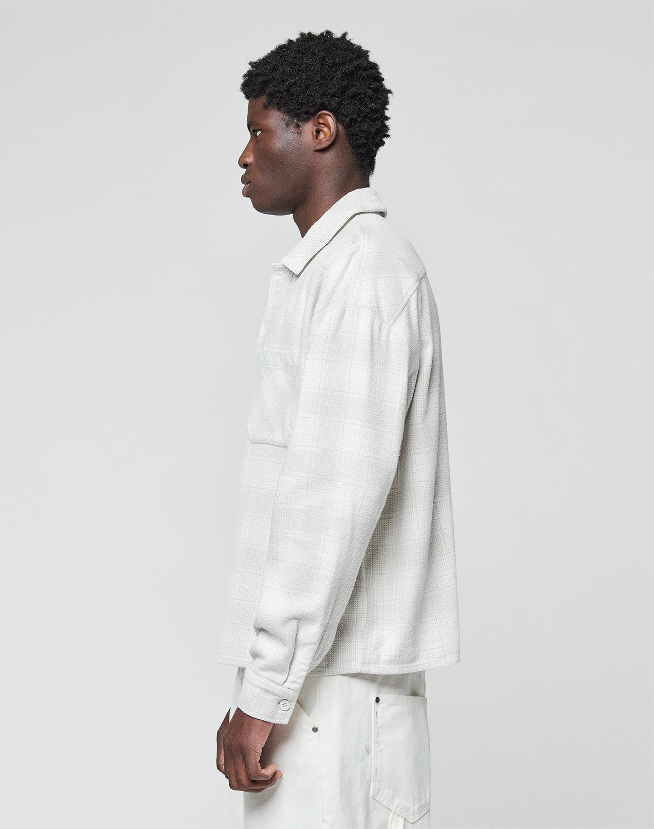 A man stands in profile against a light background, wearing a light grey LIVE FAST DIE YOUNG Vintage Flannel with a chest pocket, paired with light-colored pants.