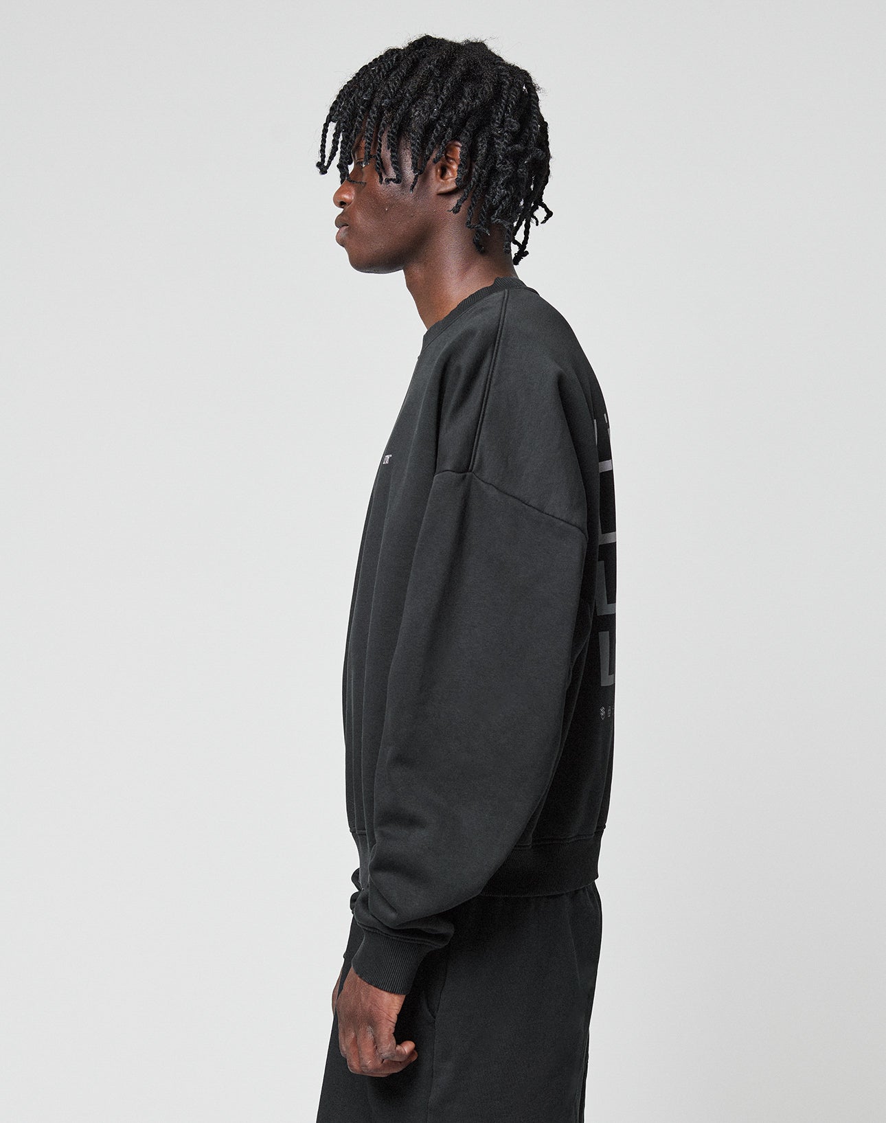 A person with short, twisted hair stands in profile facing left, wearing the LIVE FAST DIE YOUNG Basic Evidence Sweater and matching black Baumwolle Polyester pants against a plain light gray background.