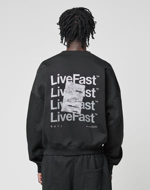 A person with short, twisted hair faces away from the camera, wearing the LIVE FAST DIE YOUNG Basic Evidence Sweater—an oversized black Baumwolle Polyester sweatshirt with bold LiveFast front and back prints layered in white graphics.