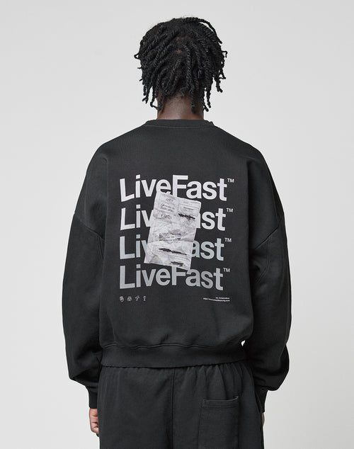 A person with short, twisted hair faces away from the camera, wearing the LIVE FAST DIE YOUNG Basic Evidence Sweater—an oversized black Baumwolle Polyester sweatshirt with bold LiveFast front and back prints layered in white graphics.