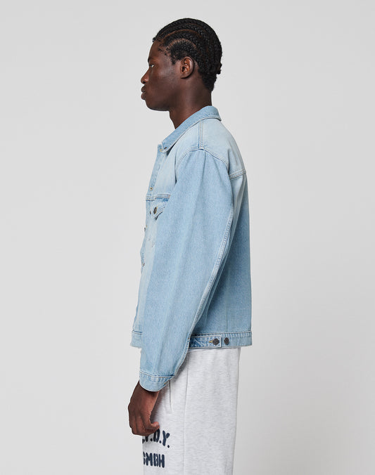 A person with short, braided hair stands in profile against a plain background, wearing the LIVE FAST DIE YOUNG LFDY Trucker Jacket in light blue denim and light gray Regular Fit sweatpants with black text on the front.