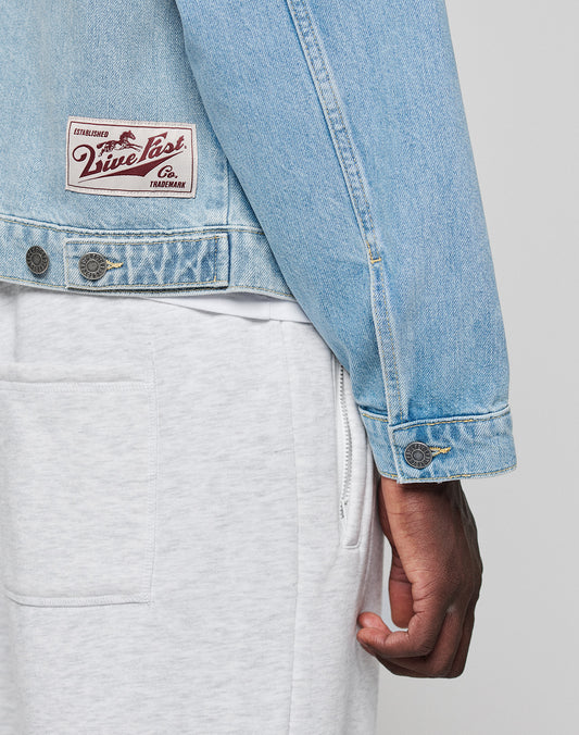 A person wearing a light blue LFDY Trucker Jacket by LIVE FAST DIE YOUNG and light gray sweatpants in regular fit, with part of their arm and hand visible.
