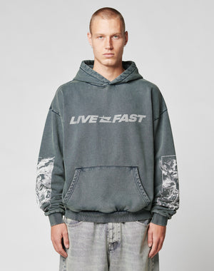 A person wearing the LFDY Window Hooded by LIVE FAST DIE YOUNG, a dark gray oversized hoodie made of 100% Baumwolle with a reflektierender LIVE FAST Frontprint and sleeve graphics, stands against a plain light gray background.
