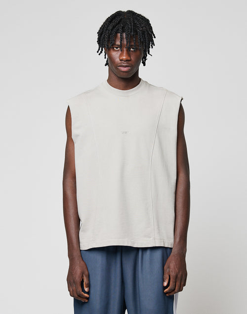 A man with short braids wears the LIVE FAST DIE YOUNG Heavy Muscle Tank in light beige Oversize Fit and loose, dark blue pants, standing facing the camera against a plain light gray background.