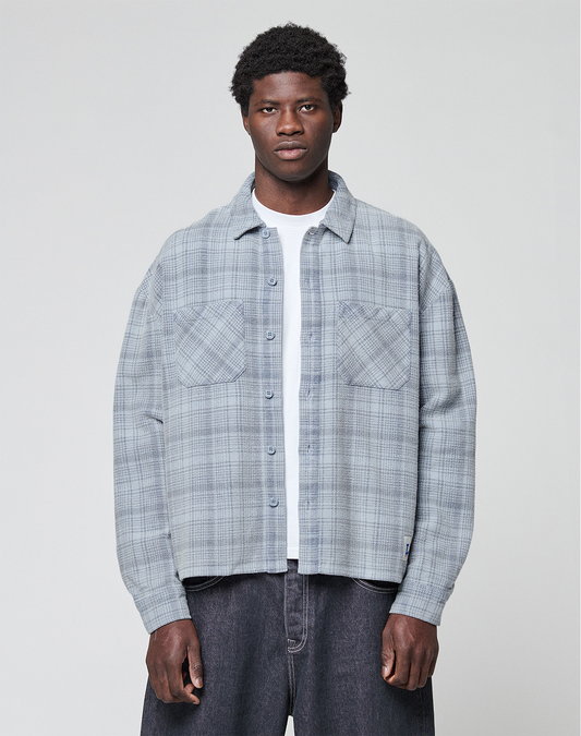 A man faces forward wearing a light gray plaid Vintage Flannel by LIVE FAST DIE YOUNG, styled in an oversize fit over a white t-shirt and dark jeans, set against a plain light background.