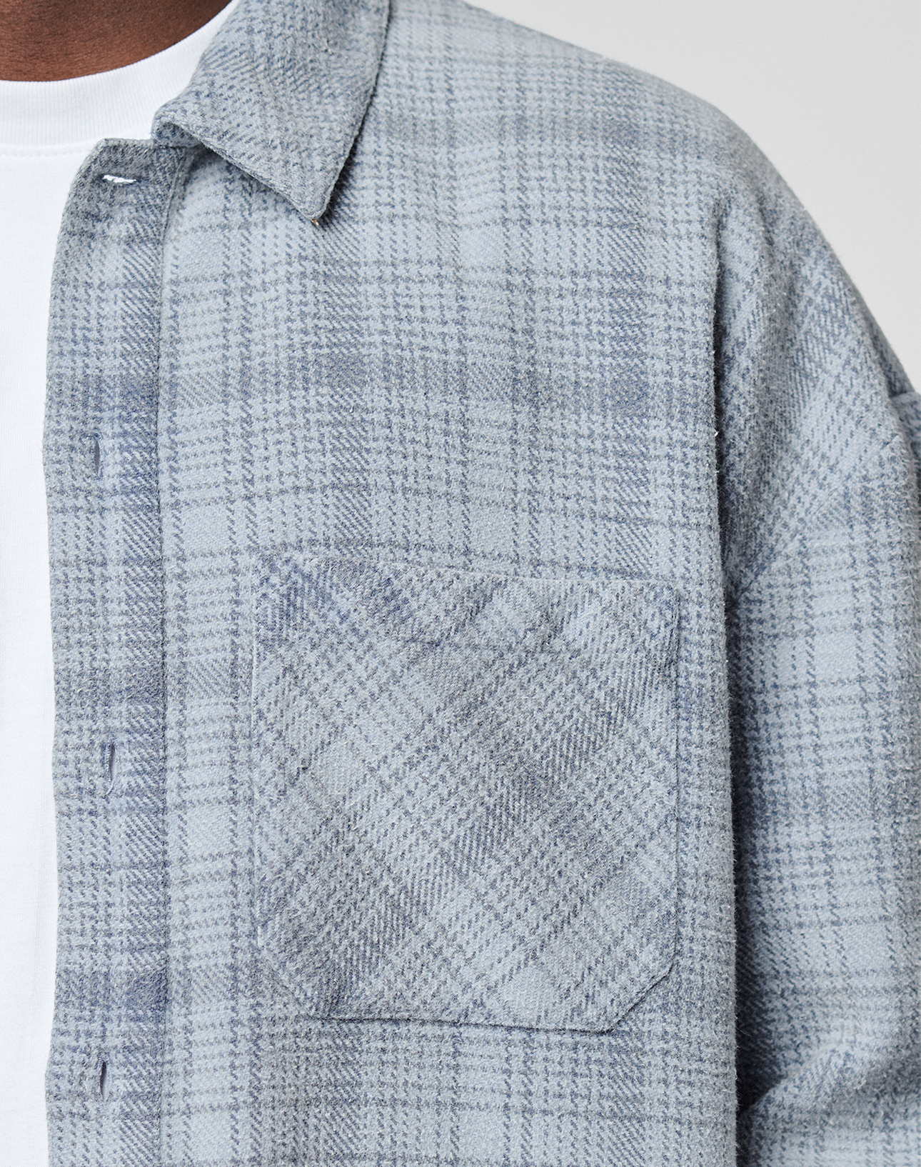 A close-up of a person wearing LIVE FAST DIE YOUNG’s Vintage Flannel in light blue and gray plaid over a white T-shirt, highlighting the front pocket, collar, and shoulder.
