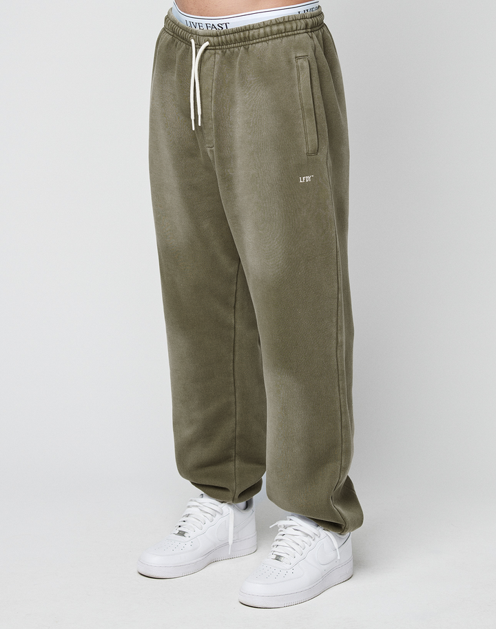 A person wears the Basic Loose Jogger by LIVE FAST DIE YOUNG in olive green with a drawstring waist, white LIVE text on the left thigh, and an elastic waistband with LIVE FAST text, styled with white sneakers on a plain white background.