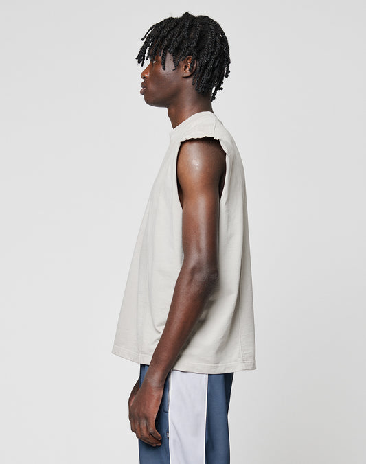 A person with short, twisted hair stands in profile wearing the LIVE FAST DIE YOUNG Heavy Muscle Tank, a sleeveless, loose-fitting beige top, and dark pants with a white side stripe against a plain light gray background.
