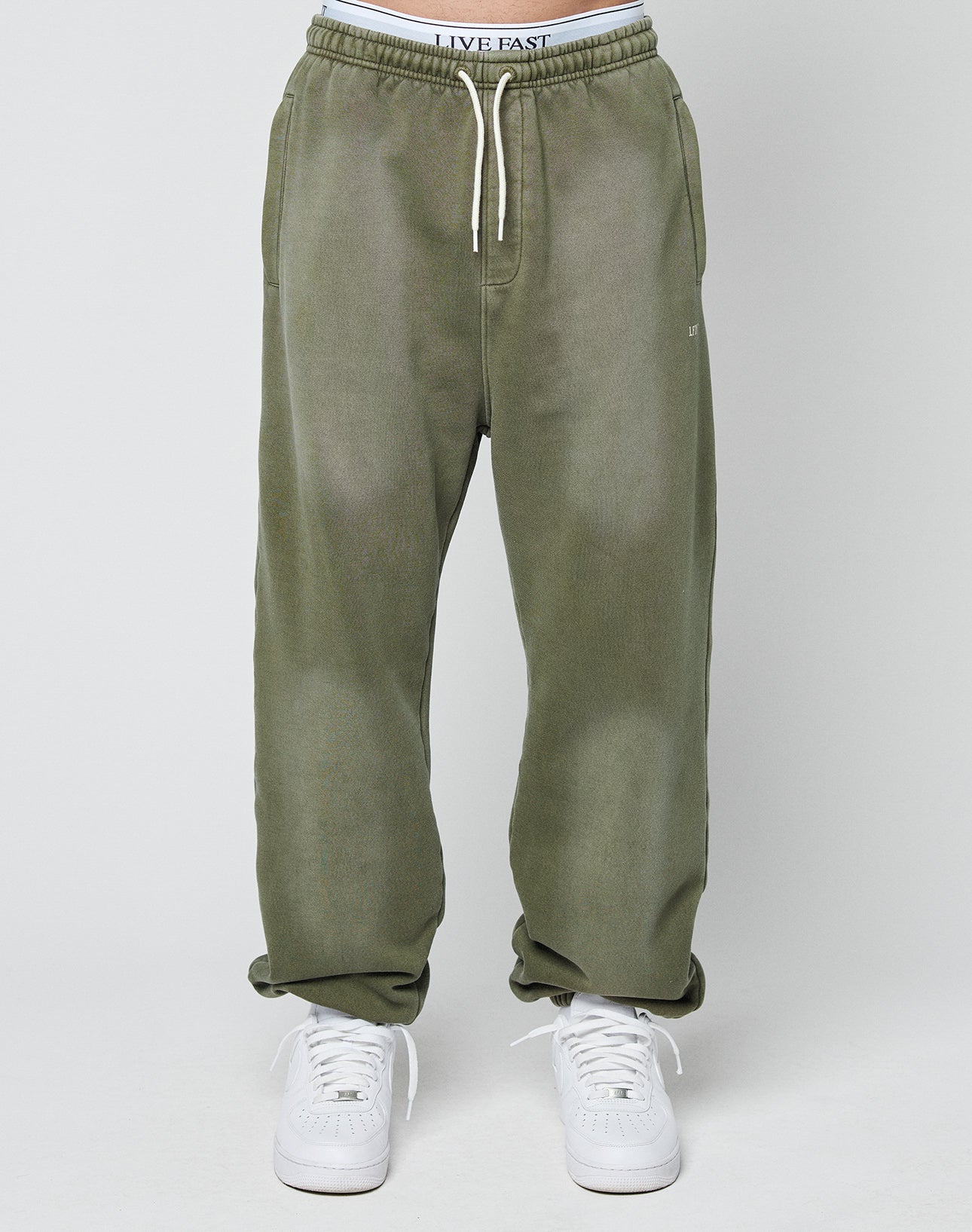 A person wearing LIVE FAST DIE YOUNG’s Basic Loose Jogger in olive green with a visible logo waistband and white sneakers stands against a plain light background.