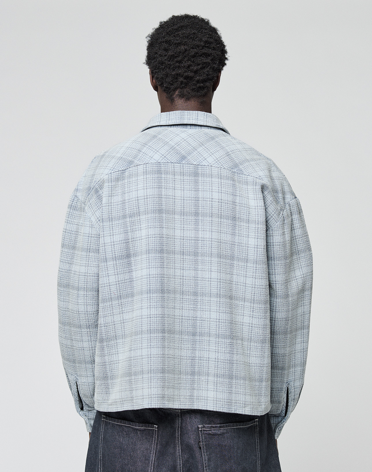 A person with short curly hair is seen from behind, wearing a light blue and gray plaid Vintage Flannel by LIVE FAST DIE YOUNG in an oversize fit with dark pants, standing against a plain light background.