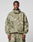 A man stands facing forward in the LIVE FAST DIE YOUNG Live Fast Nylon Jacket, featuring a green and beige camo pattern, oversize fit, front zipper, two large chest pockets, a hood with elastic drawcord, and matching pants.