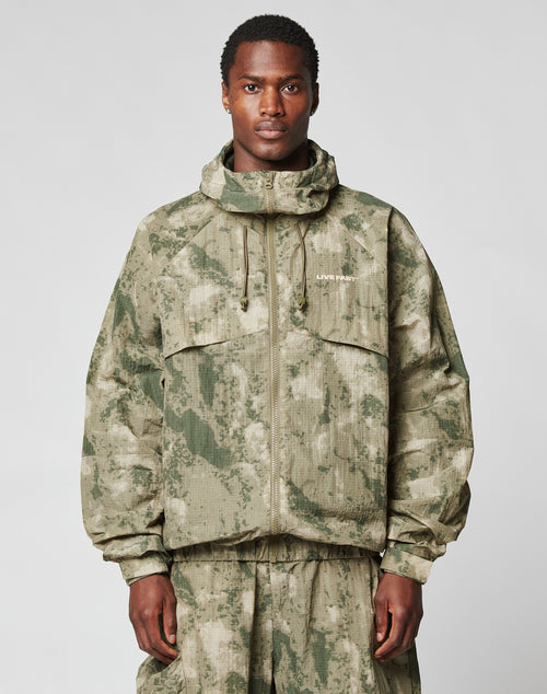 A man stands facing forward in the LIVE FAST DIE YOUNG Live Fast Nylon Jacket, featuring a green and beige camo pattern, oversize fit, front zipper, two large chest pockets, a hood with elastic drawcord, and matching pants.
