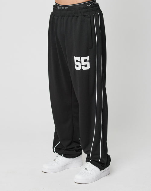 A person in LIVE FAST DIE YOUNG LFDY 55 Trackpants—black polyester with white piping, wide fit, Elastik-Bund waistband, and number 55 on the left thigh—paired with white sneakers.