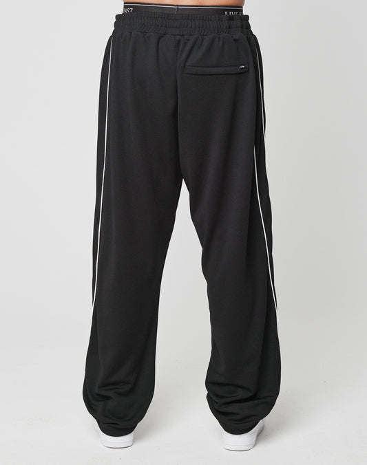 A person wears LFDY 55 Trackpants by LIVE FAST DIE YOUNG—loose-fitting black polyester pants with white side piping, Elastik-Bund waistband, wide fit, and a back pocket—styled with white shoes against a plain light backdrop.