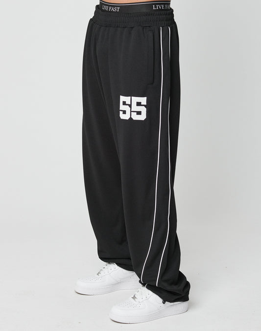 A person wears LFDY 55 Trackpants by LIVE FAST DIE YOUNG—wide fit, black with white piping, 55 printed on the left thigh, and an elastic waistband reading LIVE FAST—paired with white sneakers against a plain white background.