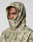 A person wears the Live Fast Nylon Jacket by LIVE FAST DIE YOUNG, an oversized green and beige camo hooded jacket with elastic drawstring, hood up and zip covering the mouth, revealing only their eyes, nose, and part of the forehead.
