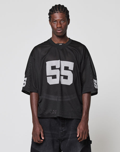 A person with braided hair wears an oversize black LFDY Mesh Jersey by LIVE FAST DIE YOUNG, featuring large white number 55 on the front and sleeves, standing against a plain light gray background.