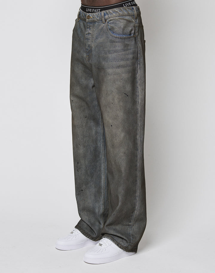 A person wearing LFDY Coated Baggy Denim by LIVE FAST DIE YOUNG, featuring a baggy fit, wide straight leg, faded black finish, minor distressing, and a branded waistband with white sneakers stands against a plain white background. | model