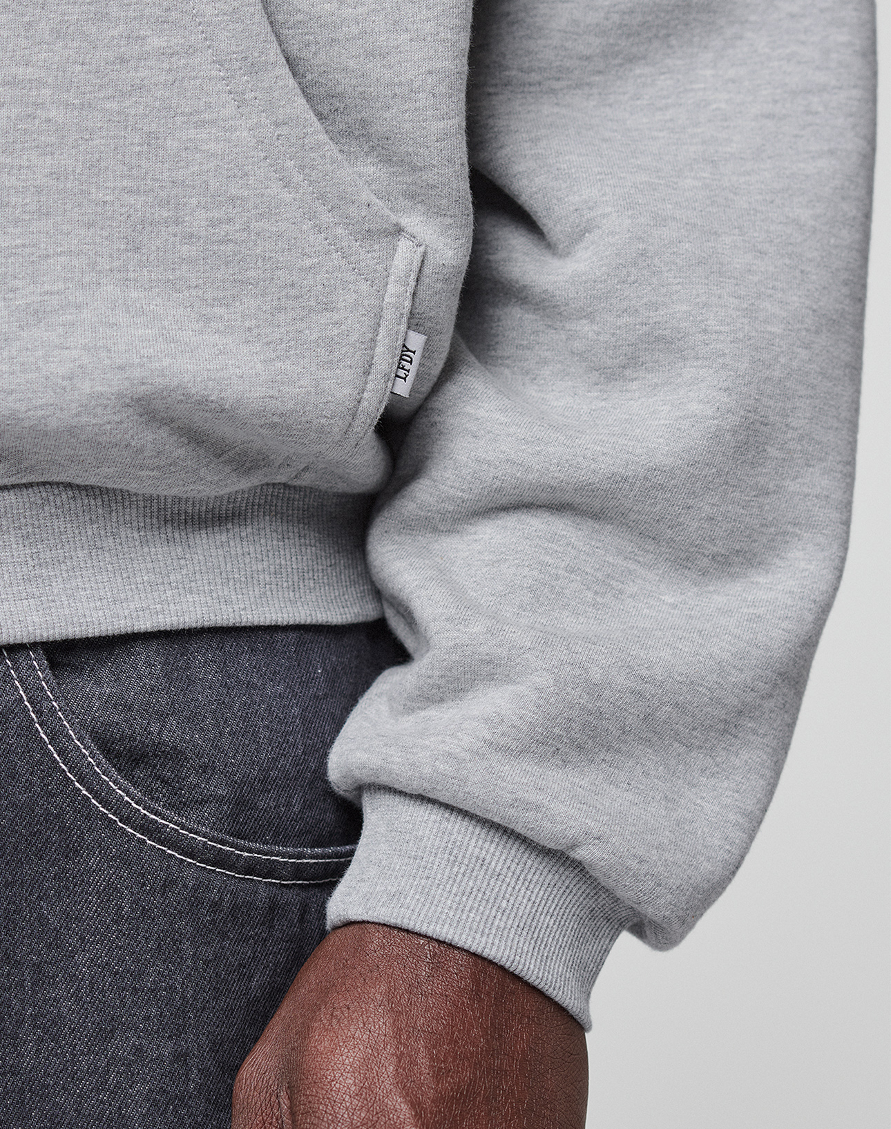 A close-up of a person wearing a light gray LIVE FAST DIE YOUNG Flannel Lined Hooded Jacket and dark denim jeans, with a small white tag near the hem and their hand visible by the pocket.