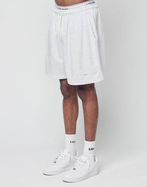 Someone wearing LIVE FAST DIE YOUNG Basic Sweat Shorts in light gray with an elastic waistband and subtle logo print, paired with white socks and sneakers, stands against a plain white background. Only their lower body is visible.
