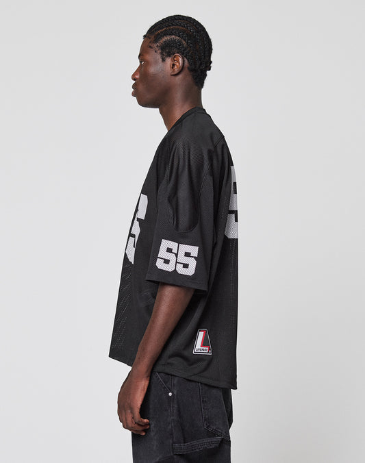 A person with braided hair stands in profile wearing the LIVE FAST DIE YOUNG LFDY Mesh Jersey—an oversized black sports jersey featuring the number 55 in white on the sleeve and side—with dark pants, against a plain light grey background.