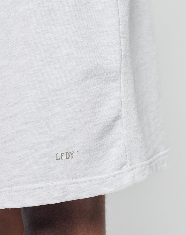 Close-up of the lower part of LIVE FAST DIE YOUNGs Basic Sweat Shorts in light gray, featuring a Logo Print and LFDY™ near the hem. Part of a dark-skinned leg appears at the bottom left against a plain, neutral background.