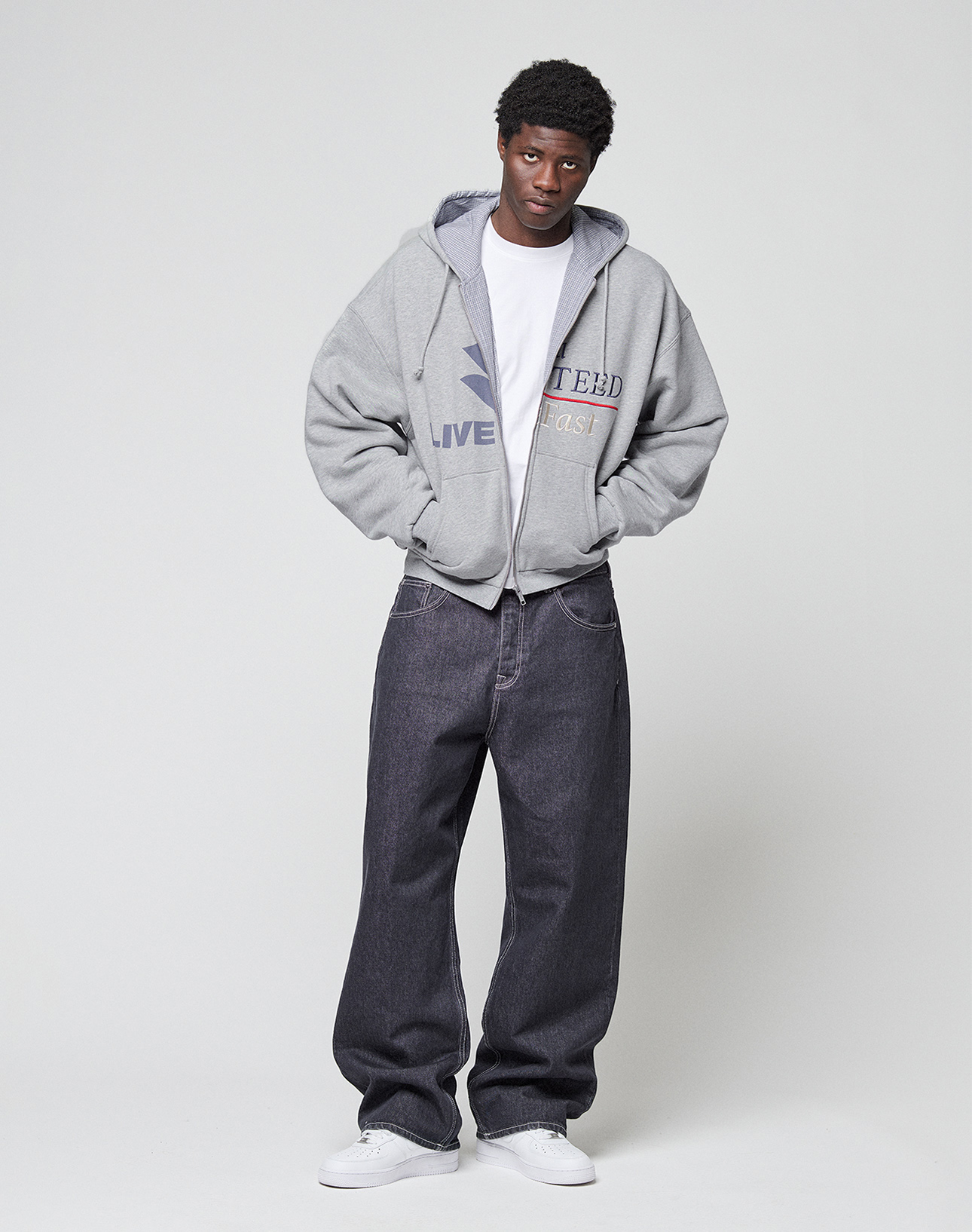 A man stands against a plain background wearing a LIVE FAST DIE YOUNG Flannel Lined Hooded Jacket over a white T-shirt, loose-fitting dark jeans, and white sneakers, hands in his pockets.