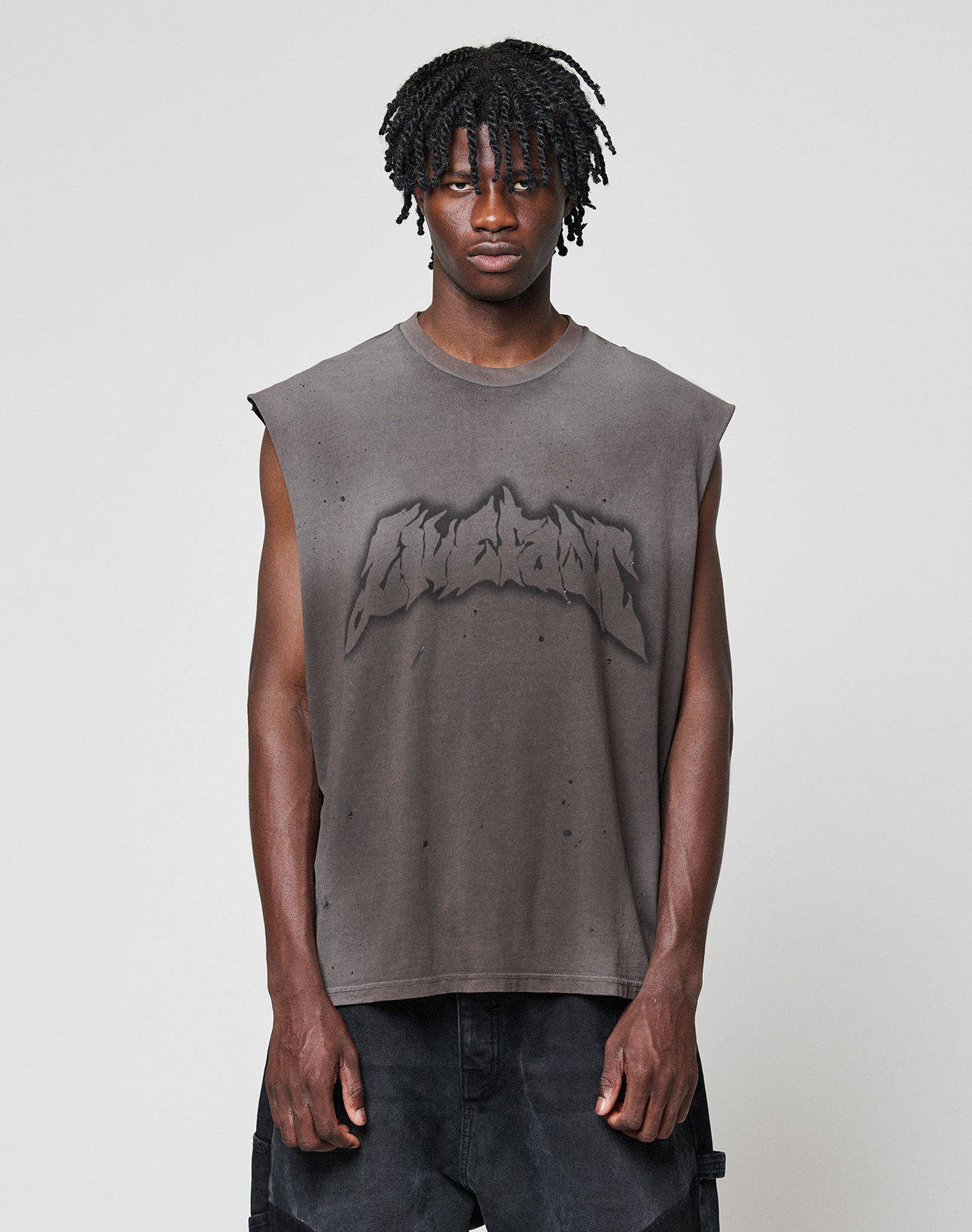 A young man with short dreadlocks wears the LFDY Flames Muscle Shirt by LIVE FAST DIE YOUNG—an oversized, sleeveless gray top with bold graphic logo and handgefertigtem Sprayeffekt—paired with black pants against a plain light gray background. | model