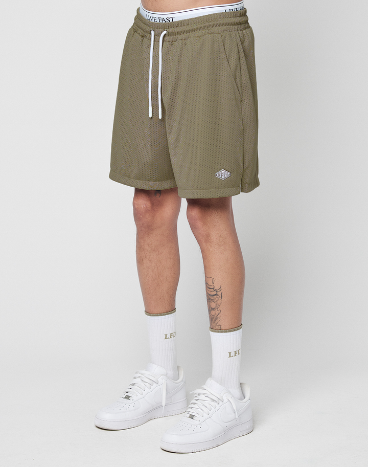Wearing LIVE FAST DIE YOUNG Basic Mesh Shorts in olive green with an elastic waistband and logo patch, paired with a white LIVE FAST waistband, white socks, and sneakers. Only the lower half is shown against a plain white background.