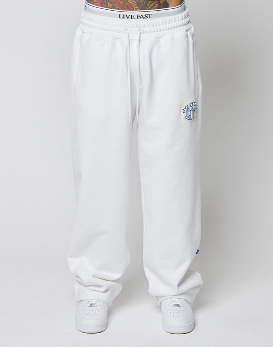 A person wears LFDY Radiator Sweatpants by LIVE FAST DIE YOUNG—oversized, white with elastic waistband and a small blue embroidered logo on the left thigh—paired with white sneakers. Tattoos show above the waistband against a plain white background.