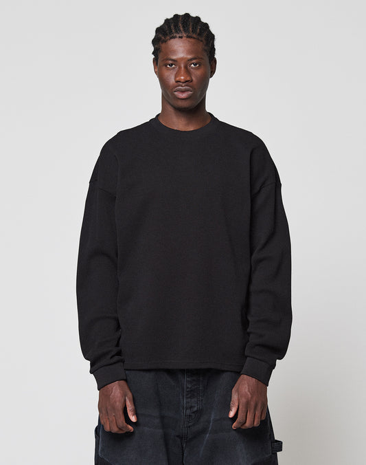 A person faces forward against a plain light gray background, wearing the LIVE FAST DIE YOUNG NY-FW Waffle Longsleeve in black with an Oversize Fit and dark pants. They have short braided hair and a neutral expression.