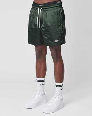 A person wears LIVE FAST DIE YOUNG Basic Satin Shorts in dark green with a white drawstring and elastic waistband, paired with white crew socks featuring green stripes and white sneakers, standing against a plain white background.