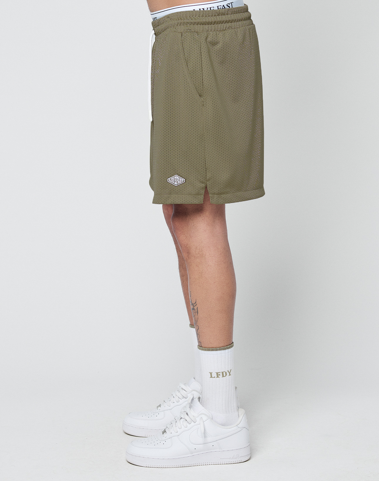 A person wearing olive green LIVE FAST DIE YOUNG Basic Mesh Shorts with a logo patch and elastic waistband, white crew socks with LFDY text, and white Nike sneakers stands sideways against a plain white background.