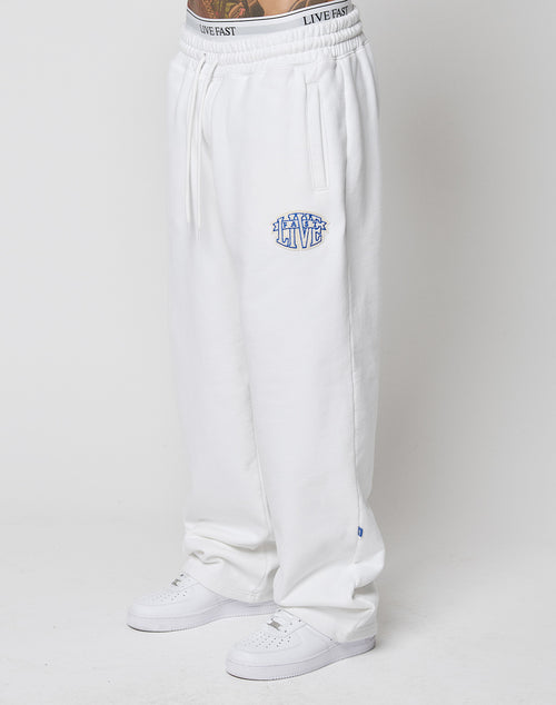 Model wears loose white LFDY Radiator Sweatpants by LIVE FAST DIE YOUNG, featuring an elastic waistband with LIVE FAST text and a blue-and-white chenille LIVE patch on the left thigh. Styled with white sneakers; simple light background.