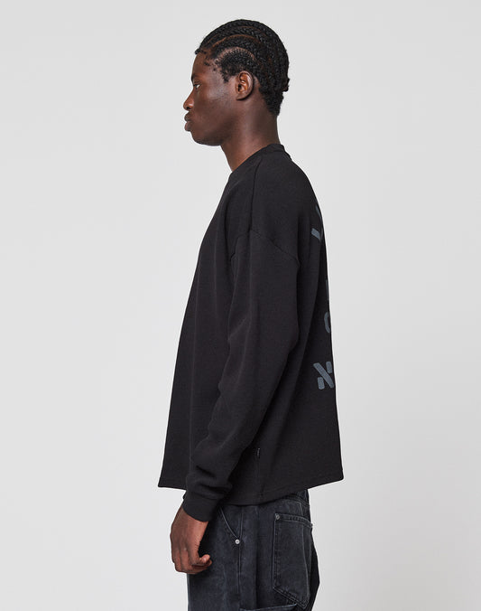 A person with braided hair stands sideways, wearing the LIVE FAST DIE YOUNG NY-FW Waffle Longsleeve in black and matching 100% Baumwolle pants, against a plain light gray background.