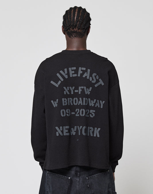 A person with braided hair, seen from behind, wears the LIVE FAST DIE YOUNG NY-FW Waffle Longsleeve in black (100% Baumwolle, oversize fit) featuring a gray backprint: LIVE FAST NY-FW W BROADWAY 09-2025 NEWYORK. Background is plain and light.