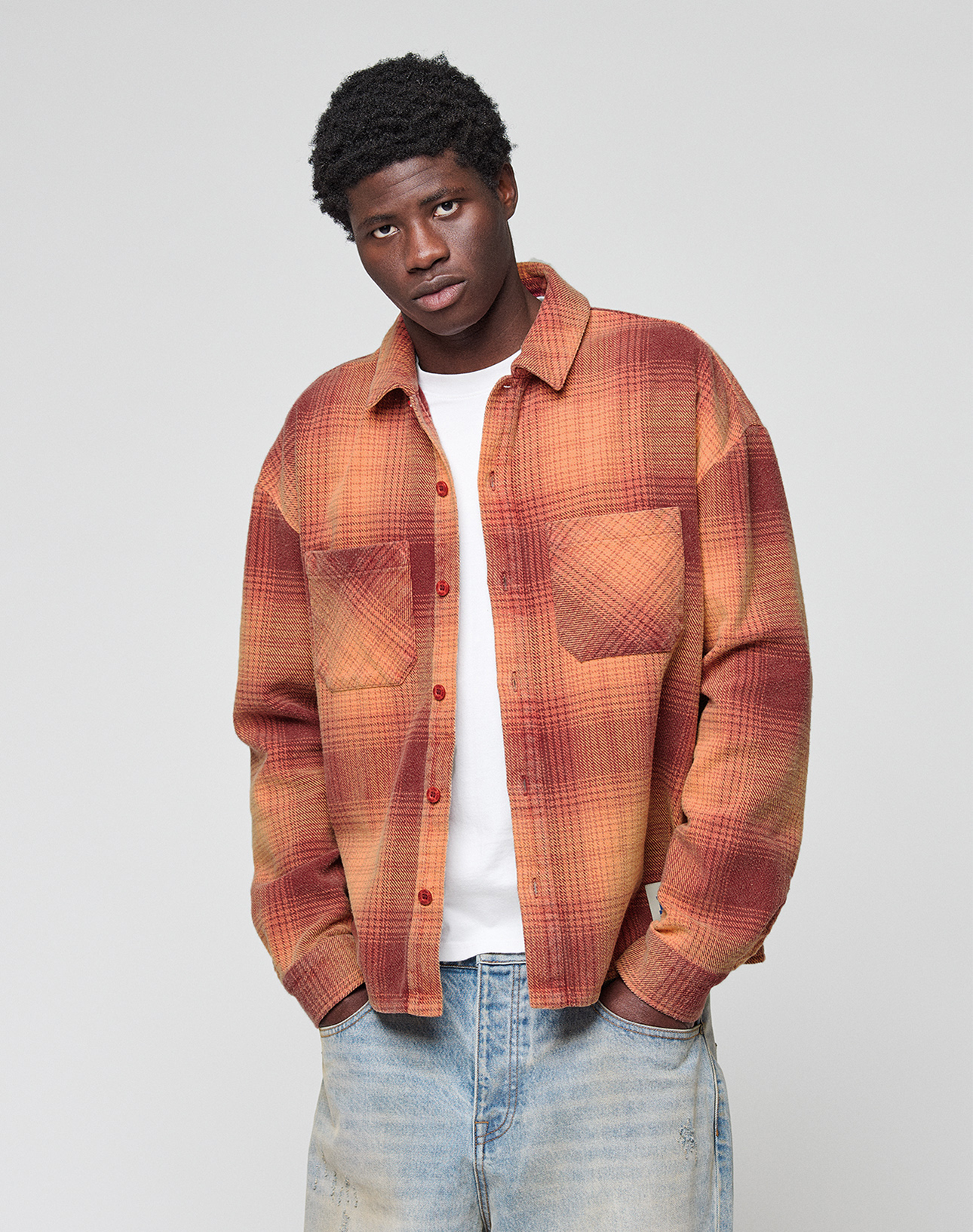 A young man in a red and orange LIVE FAST DIE YOUNG Vintage Flannel over a white t-shirt and light blue jeans stands with hands in his pockets against a plain, light gray background.