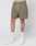A person wears olive green Basic Mesh Shorts by LIVE FAST DIE YOUNG with a white logo patch and elastic waistband, paired with white “LIVE FAST” socks and white sneakers. Their tattooed legs stand out against a plain light background.