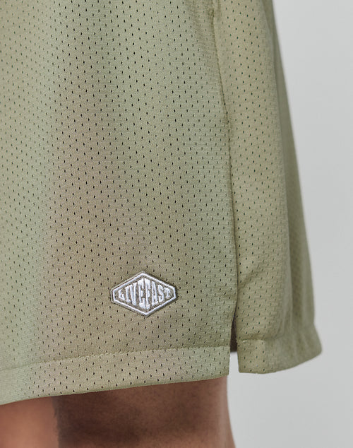 Close-up of LIVE FAST DIE YOUNG Basic Mesh Shorts in olive green polyester, featuring a small white logo patch near the hem; a portion of the wearer’s leg is visible at the bottom.