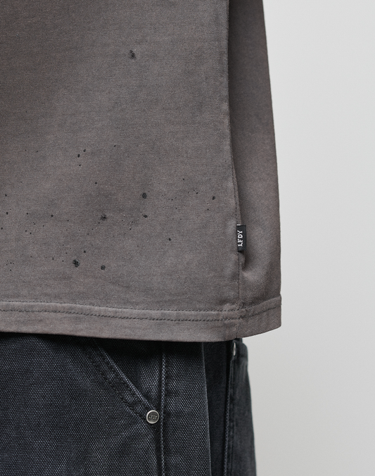 A close-up of the lower part of the gray LFDY Flames Muscle Shirt by LIVE FAST DIE YOUNG with a small black side tag, paired with dark denim. The shirt features a hand-sprayed effect and subtle black speckles near the hem.