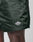 Close-up of a person wearing LIVE FAST DIE YOUNG Basic Satin Shorts in dark green, featuring an elastic waistband and a small white logo patch by the hem. Their dark skin stands out against the light background for a crisp, modern effect.