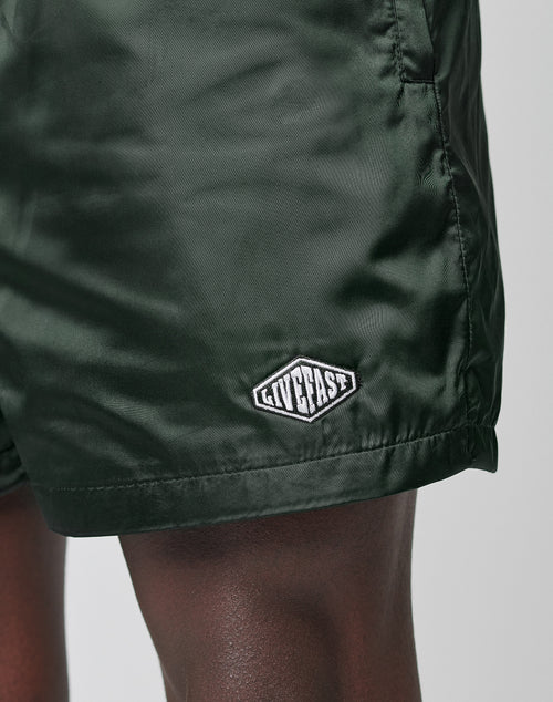 Close-up of a person wearing LIVE FAST DIE YOUNG Basic Satin Shorts in dark green, featuring an elastic waistband and a small white logo patch by the hem. Their dark skin stands out against the light background for a crisp, modern effect.