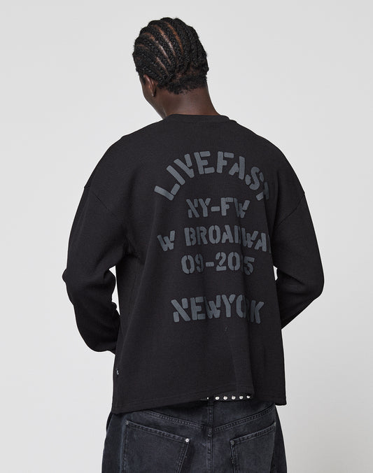 Seen from behind, a person with braided hair wears the LIVE FAST DIE YOUNG NY-FW Waffle Longsleeve—an oversize black 100% cotton sweater with gray backprint text: LIVEFAST NY-FW W BROADWAY 09-2015 NEWYORK. Plain white background.