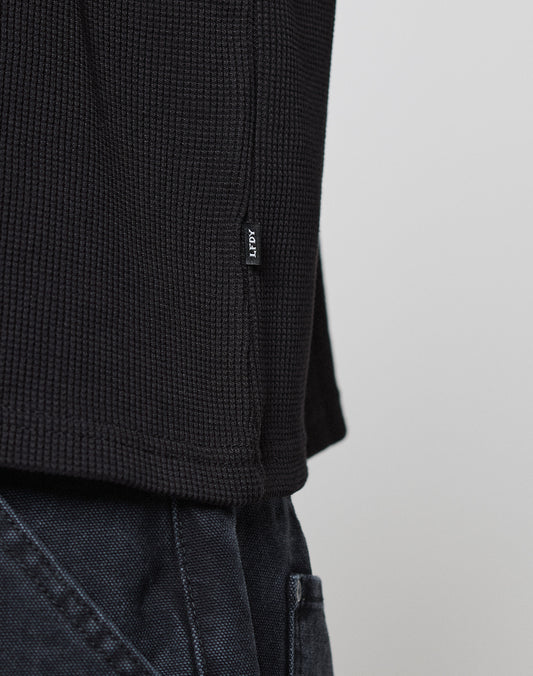 Close-up of the LIVE FAST DIE YOUNG NY-FW Waffle Longsleeve in black, featuring a textured oversize fit and a small black-and-white hem tag, styled with dark pants against a light background.
