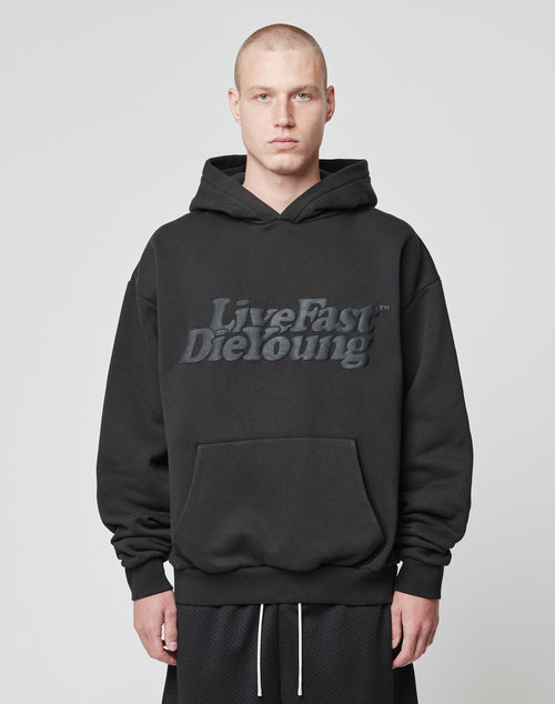 A person with a shaved head wears an oversize fit black Basic Hooded by LIVE FAST DIE YOUNG, with embossed branding on the front, standing against a plain light gray background.