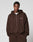 A man stands against a plain light background, hands in his pockets, wearing the LFDY Pleated Hooded Zip by LIVE FAST DIE YOUNG—an oversize fit brown zip-up hoodie aus 100% Baumwolle with a small white LIVE FAST logo and Kapuze mit Kordel.